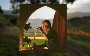 Man pouring wine from a dropper into a wine glass over a barrel with vineyard in the background